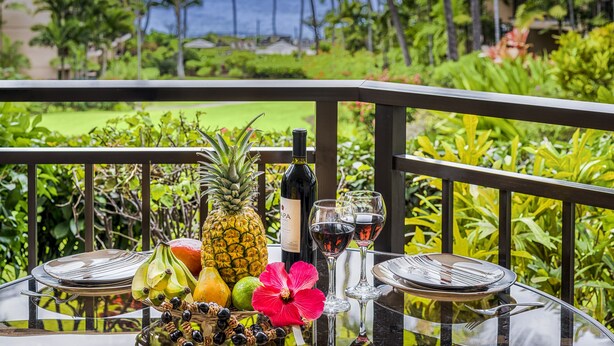 Une table dressée avec des fruits, du vin et des verres, sur un balcon offrant une vue sur l’océan et la végétation tropicale.