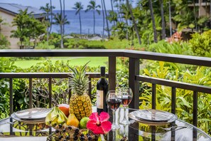 Una mesa con frutas, vino y copas en un balcón con vistas al océano y plantas tropicales.