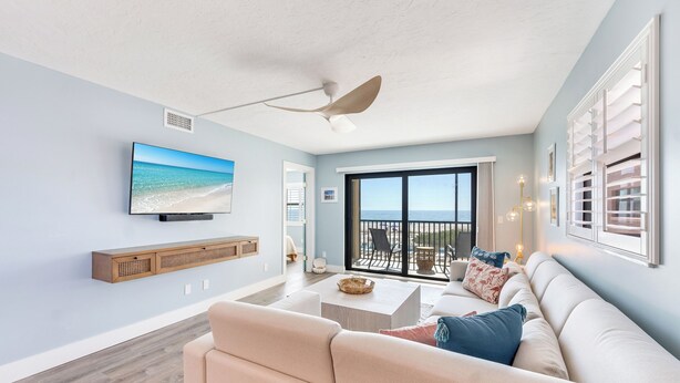 A living room with a large TV, a ceiling fan, a sofa, and a view of the ocean through a sliding glass door.