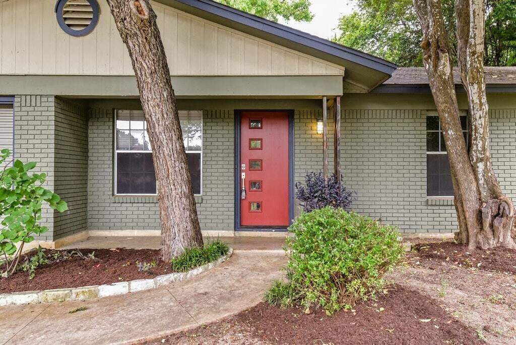 A two-story building with a red door, brick exterior, and a small front yard with a tree and shrub.