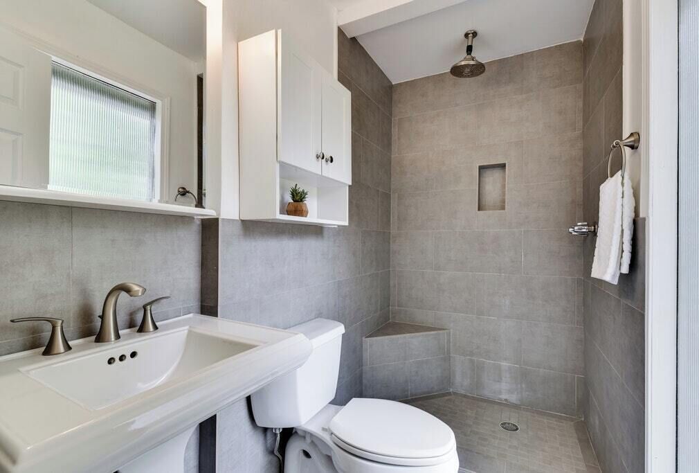 Een moderne badkamer met een wit wastafelmeubel, toilet en douche. Er is een raam met zonwering, een handdoekhouder en een kleine plant op de plank.