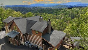 A log cabin with a stone chimney, a porch, and a view of the surrounding forest and mountains. - Mtn Views-4BR-GameRm-More! (Sevierville)