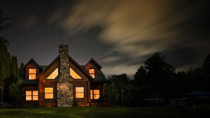 Une cabane en rondins dotée de nombreuses fenêtres et d’une cheminée en pierre, entourée d’une cour verdoyante et d’arbres.