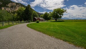 Ein Schotterweg führt zu einem roten Haus mit weißem Zaun, umgeben von grünem Gras und Bäumen, mit Bergen im Hintergrund.