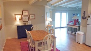 A dining area with a wooden table and white chairs, a small counter with a lamp, and framed pictures on the wall.