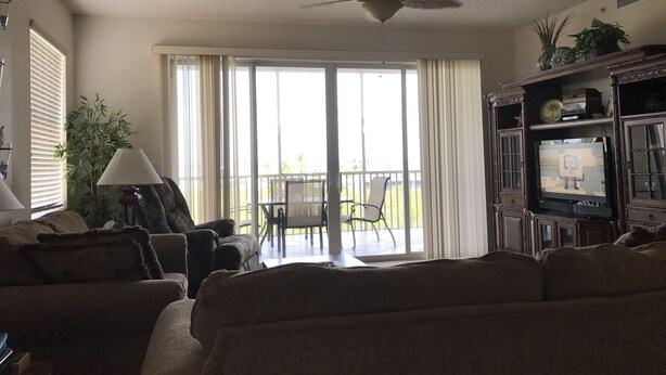 Una sala de estar con ventilador de techo, un televisor sobre un mueble de madera, un sofá, una lámpara y vistas a un balcón con sillas.