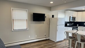 Cocina compacta con gabinetes blancos, microondas integrado, refrigerador y televisor de pantalla plana montado en la pared.