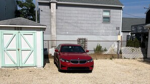 Un coche rojo estacionado en un camino de grava frente a una casa con un garaje blanco y una valla de metal.