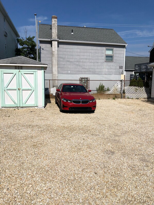 A red car parked in a gravel driveway in front of a house with a white garage and a metal fence. - 3 Bed 1 Bath (2nd Floor) (Seaside Heights)