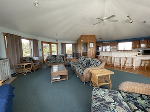Tilava olohuone, jossa on rottinkihuonekaluja, puukehikoilla varustettu keittiö ja suuret ikkunat.