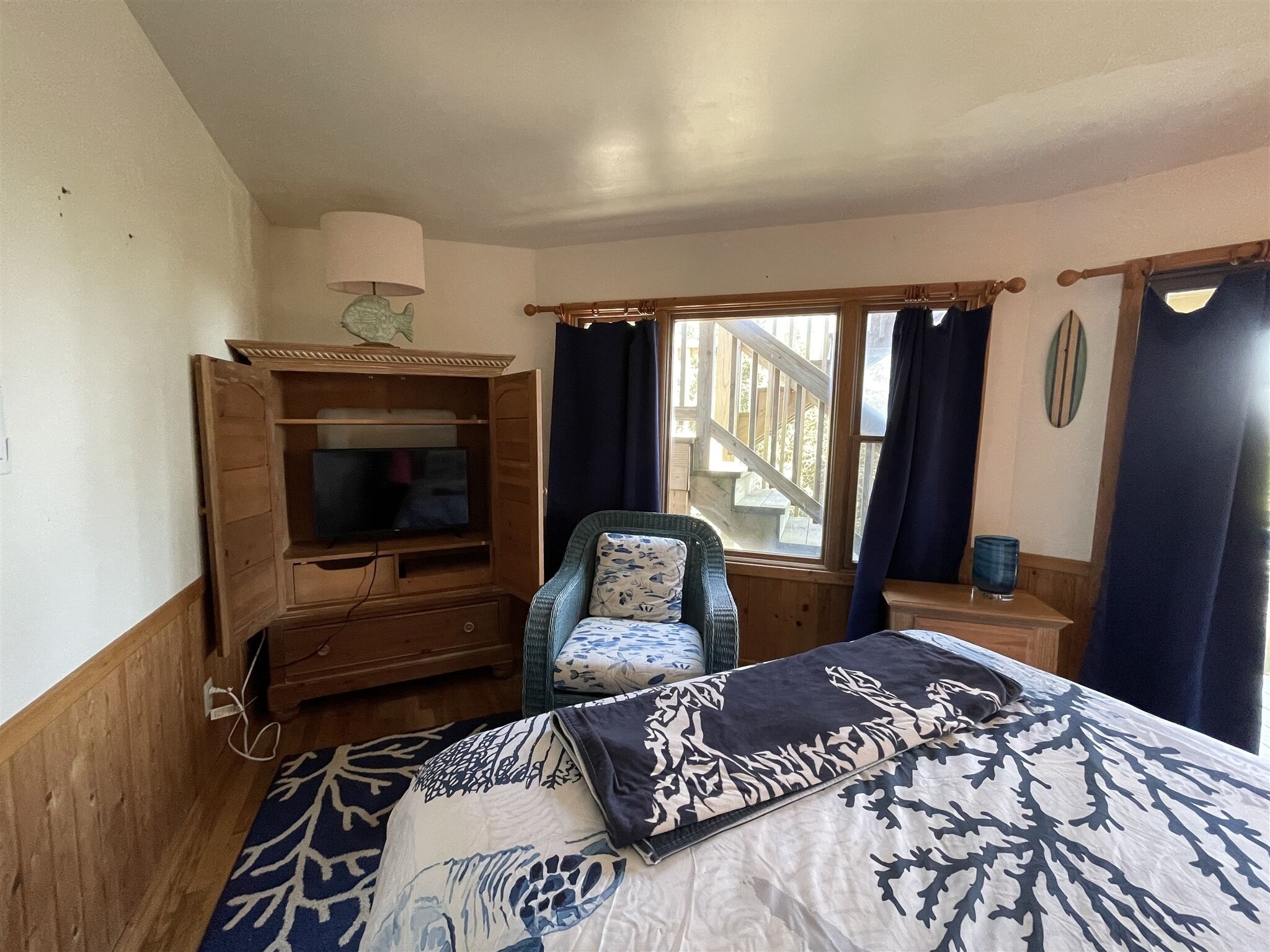 A bedroom with a bed, a television in a wooden cabinet, a chair, a window with curtains, and a view of a wooden staircase.