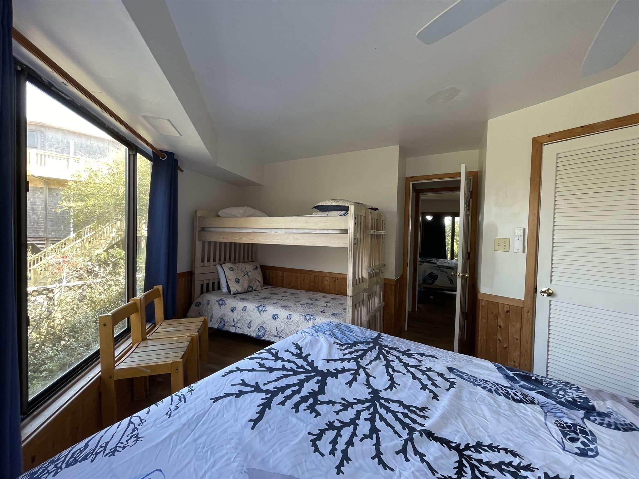 Een kamer met stapelbedden, een houten stoel, een bed met een sprei met bladprint en een raam met gordijnen.