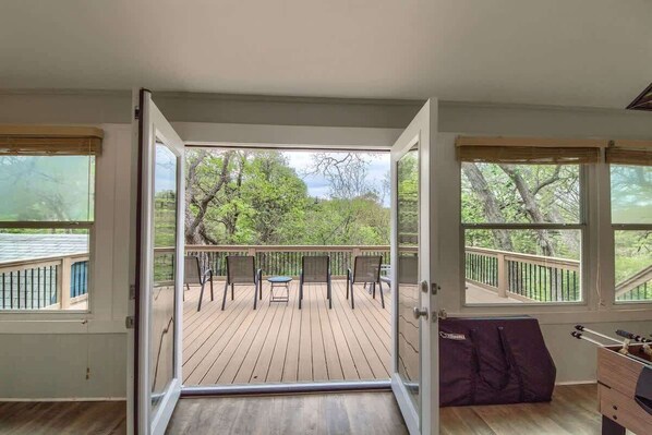Une porte ouverte donne sur une terrasse Ă©quipĂ©e dâune table et de chaises, dâun barbecue et offrant une vue sur les arbres.