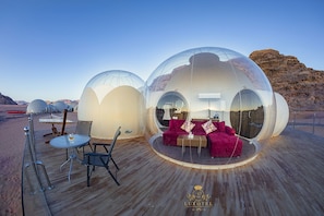 Transparent dome-shaped structures with seating and tables on a wooden deck, set against a desert backdrop.