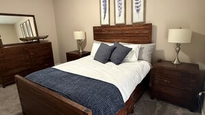 Quarto com cama de madeira, duas mesas de cabeceira com luminárias, cômoda e três quadros decorativos na parede.