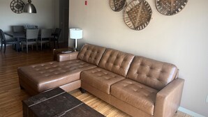 Uma sala de estar com um sofá seccional de couro marrom, uma mesa de centro, uma área de jantar com mesa e cadeiras, e três objetos decorativos fixados na parede.