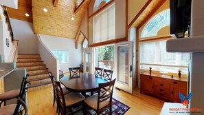 Un comedor amplio con una mesa y sillas de madera, una escalera que lleva a niveles superiores y ventanas grandes que ofrecen vistas al exterior.