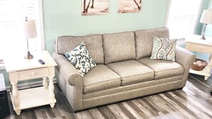 A living room with a beige sofa, two matching throw pillows, a small white side table with a lamp, and a wooden floor.