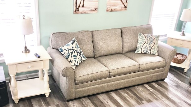 A living room with a beige sofa, two matching throw pillows, a small white side table with a lamp, and a wooden floor.
