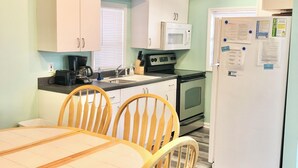 Cocina compacta con gabinetes blancos, microondas, estufa y refrigerador. Hay una mesa de comedor con sillas y un tapete a rayas.