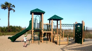 A playground with a slide and climbing structures, surrounded by sand and palm trees.