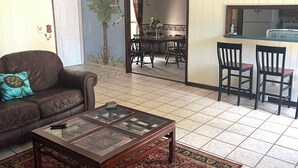A living room with a brown leather sofa, a glass-top coffee table, and a patterned rug. There is a dining area with a table and chairs visible through an open doorway.
