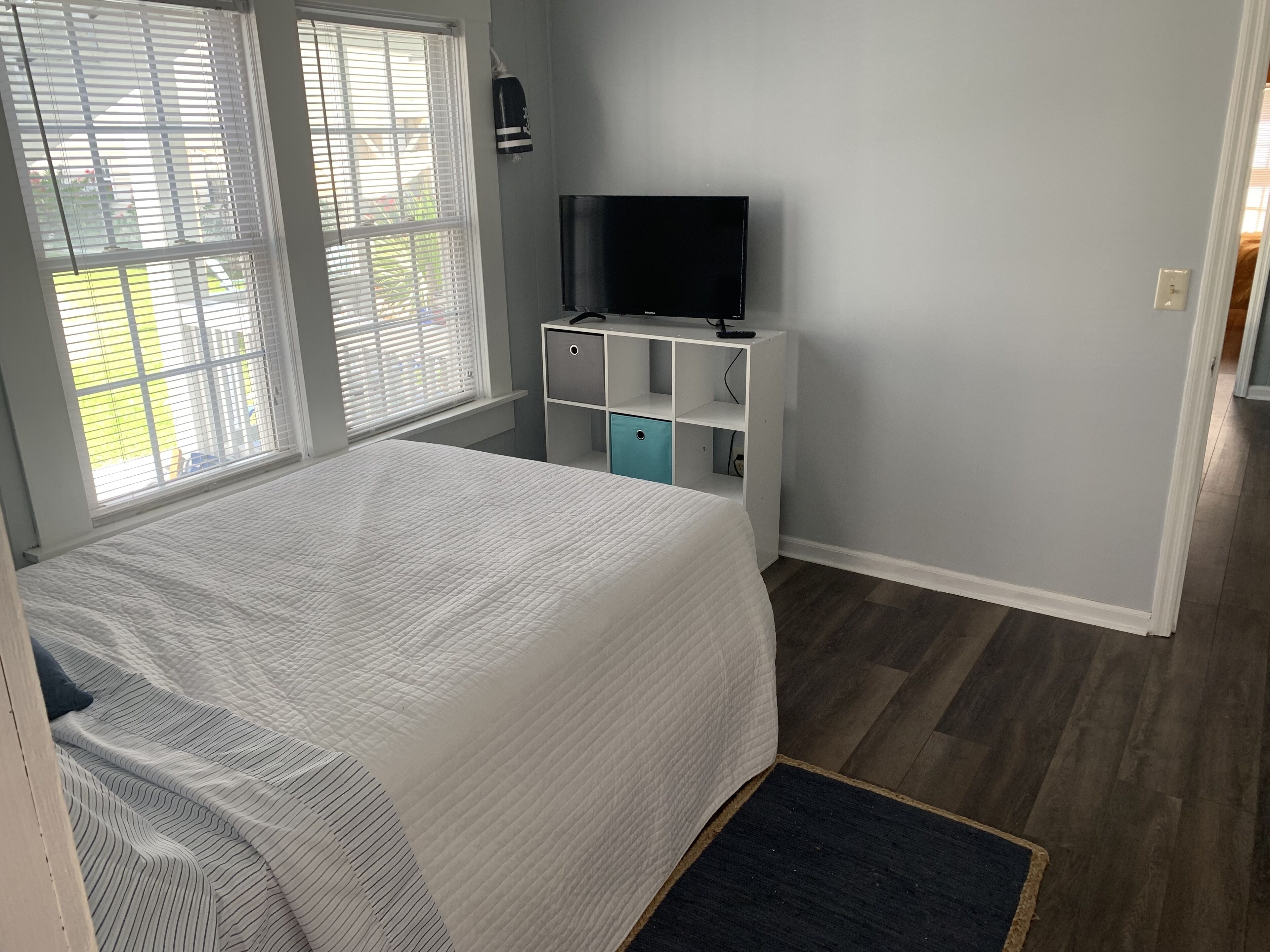 A bedroom with a bed, a TV on a white cabinet, and a window with blinds.