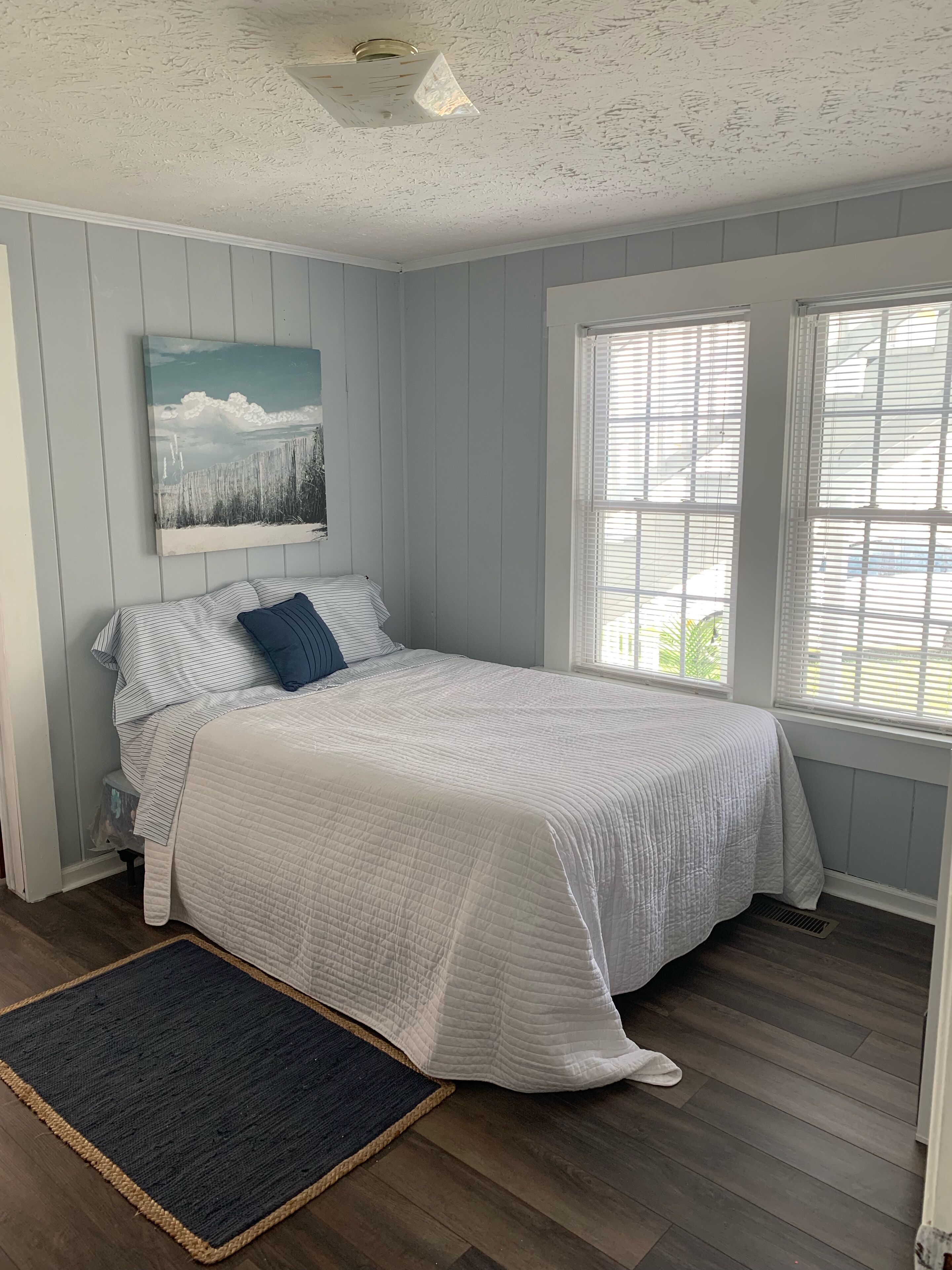 Une chambre à coucher avec un lit, une fenêtre avec des stores, un ventilateur de plafond et un tableau au mur.