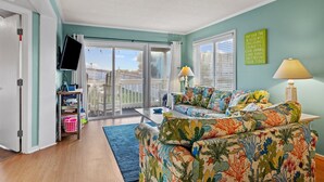 A living room with a sofa, a TV, and a window with blinds. - Perfectly pool side at APATB! King, Full, 2 Twins (Atlantic Beach)