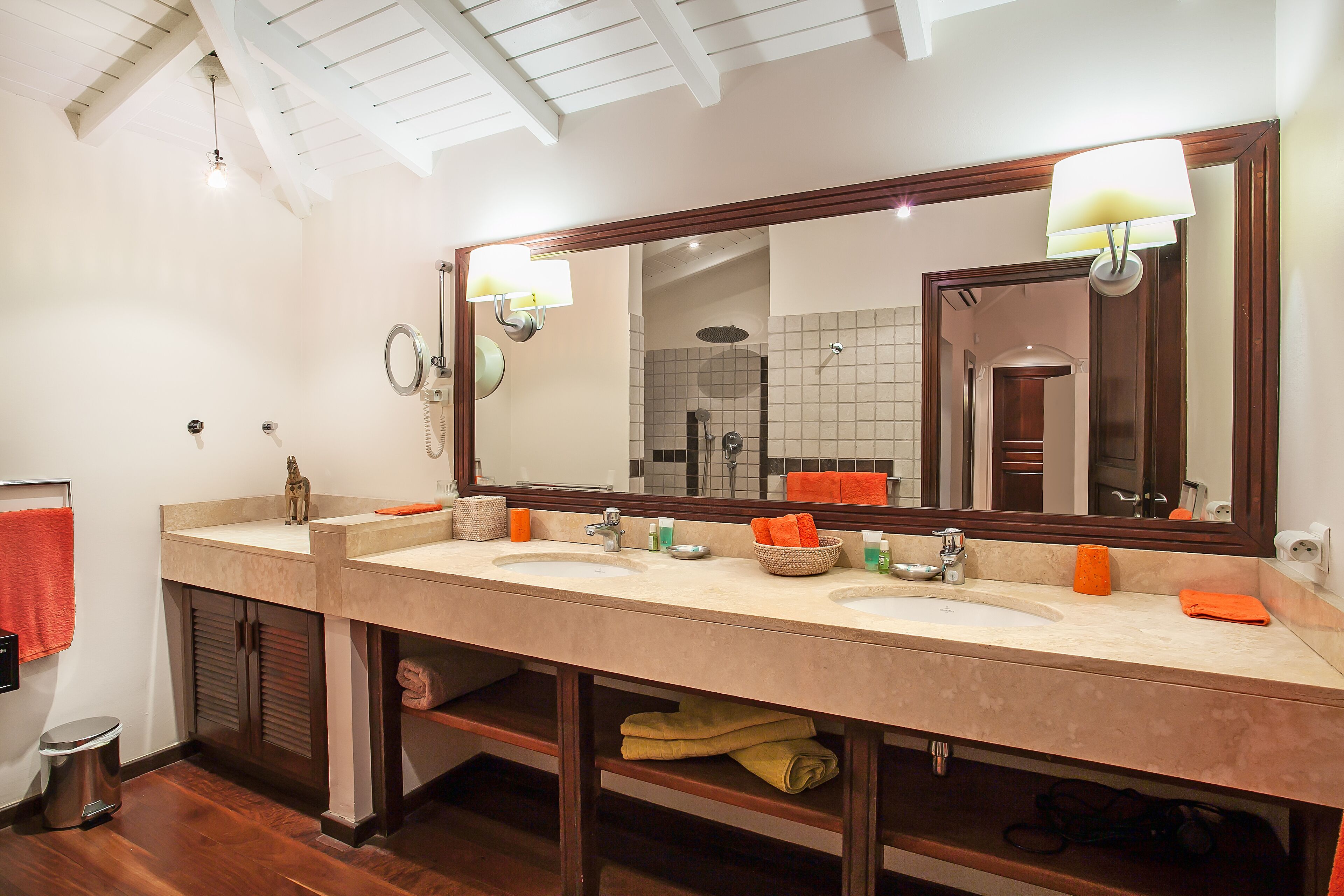 A bathroom with a large mirror, two sinks, and a shower. There are towels and toiletries on the sinks.