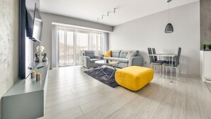 A modern living room with a grey sofa, yellow ottoman, and a dining area with a table and chairs.