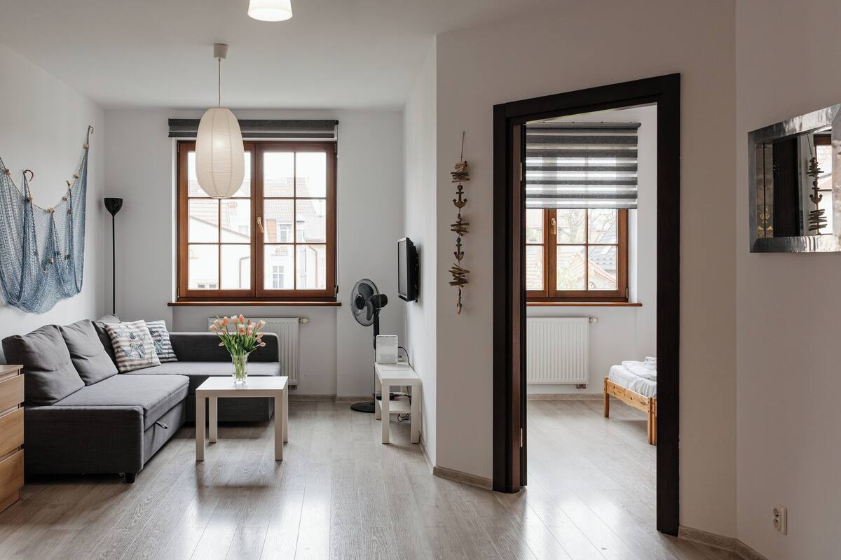 Una sala de estar moderna con sofá, una mesita de centro y televisor. Hay dos ventanas con persianas, una lámpara colgante y piso de madera.