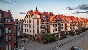 A row of traditional European-style buildings with red-tiled roofs and balconies, a cobblestone street, and a clear sky.