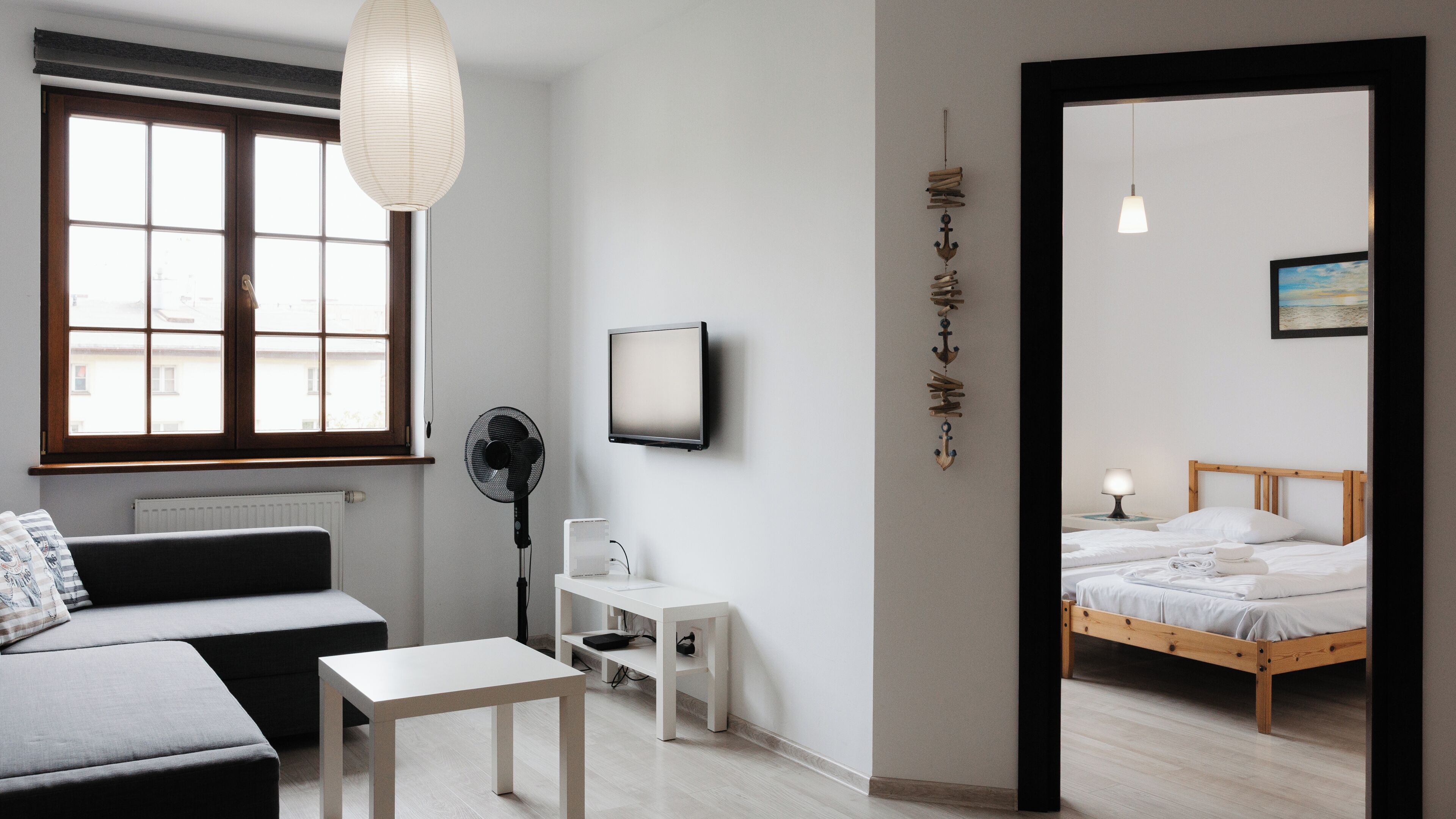 A modern hotel room with a sofa, a small table, a desk, a bed, a TV, and a wall-mounted fan.