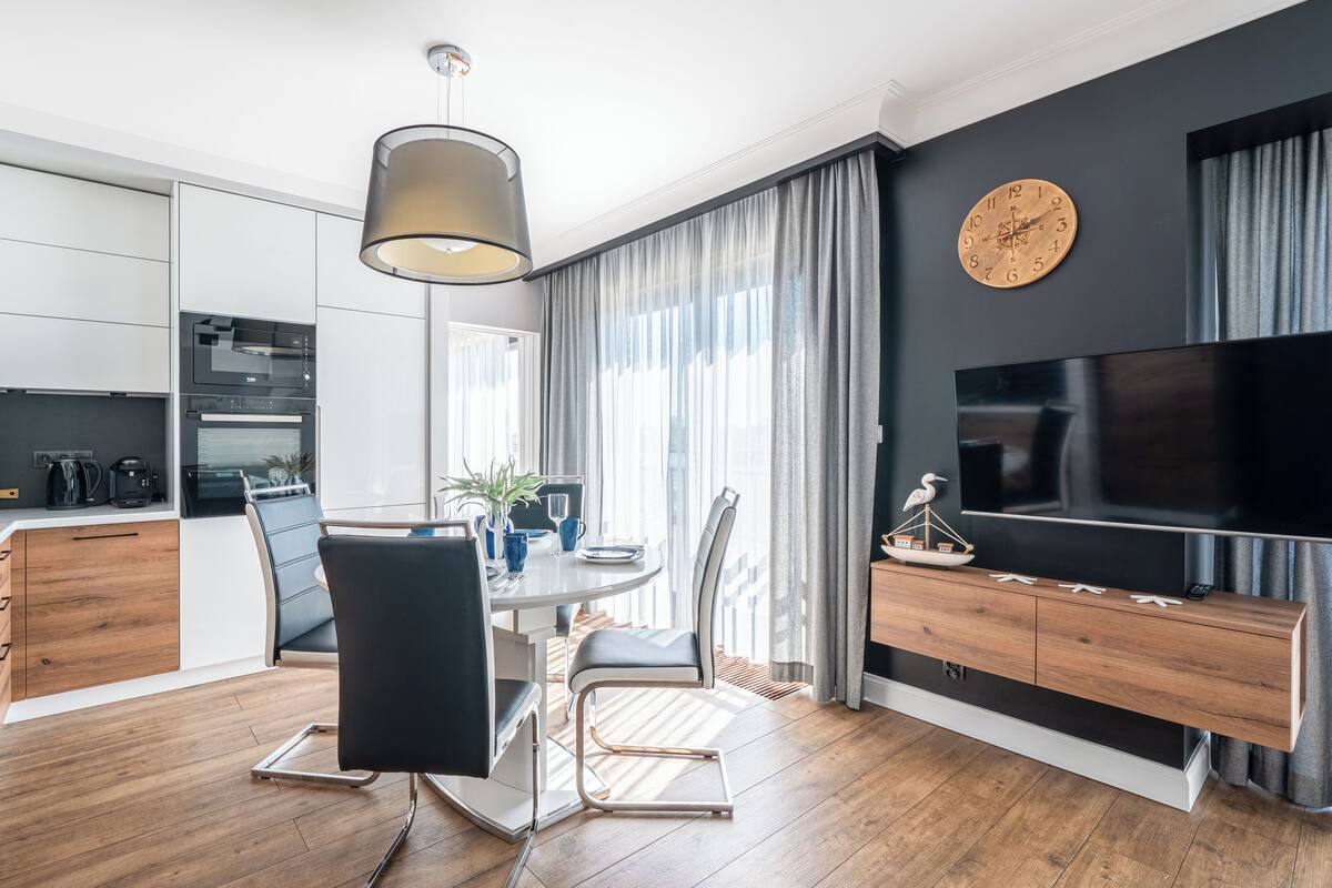 Una cocina moderna con electrodomésticos integrados, un comedor con mesa y sillas, un televisor montado en la pared y un amplio ventanal con cortinas.