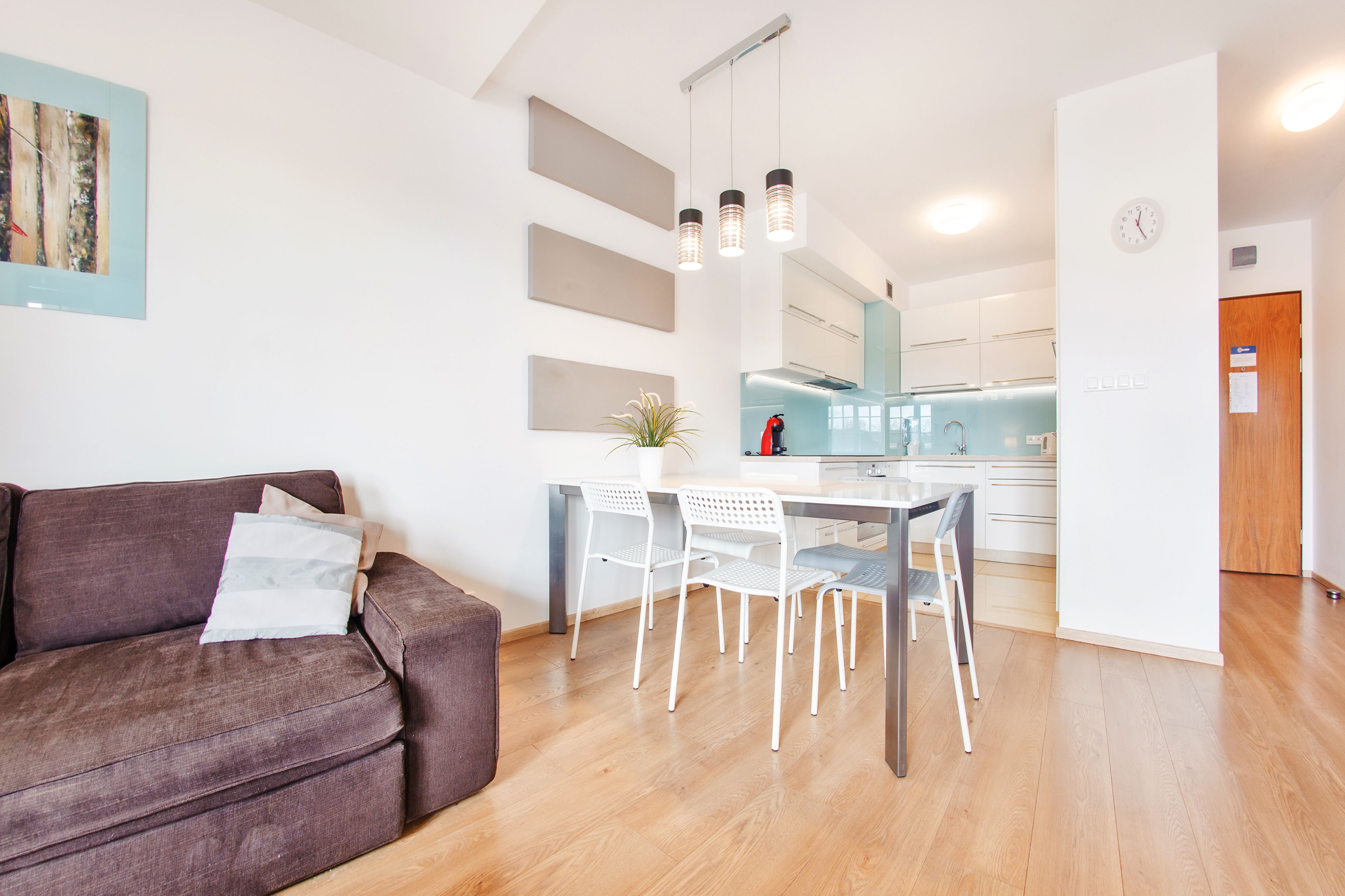 A modern living room with a brown sofa, a dining table with white chairs, and a kitchen in the background.