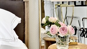 Una cama bien hecha con cabecera, una mesita de noche con un jarrón de flores y un espejo que refleja un baño con un perchero.
