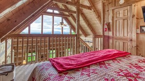 A wooden cabin bedroom with a sloped ceiling, a bed with a red blanket, a ceiling fan, and a view of the outdoors. - View-topia! VIEWS, VIEWS AND MORE VIEWS (Sevierville)