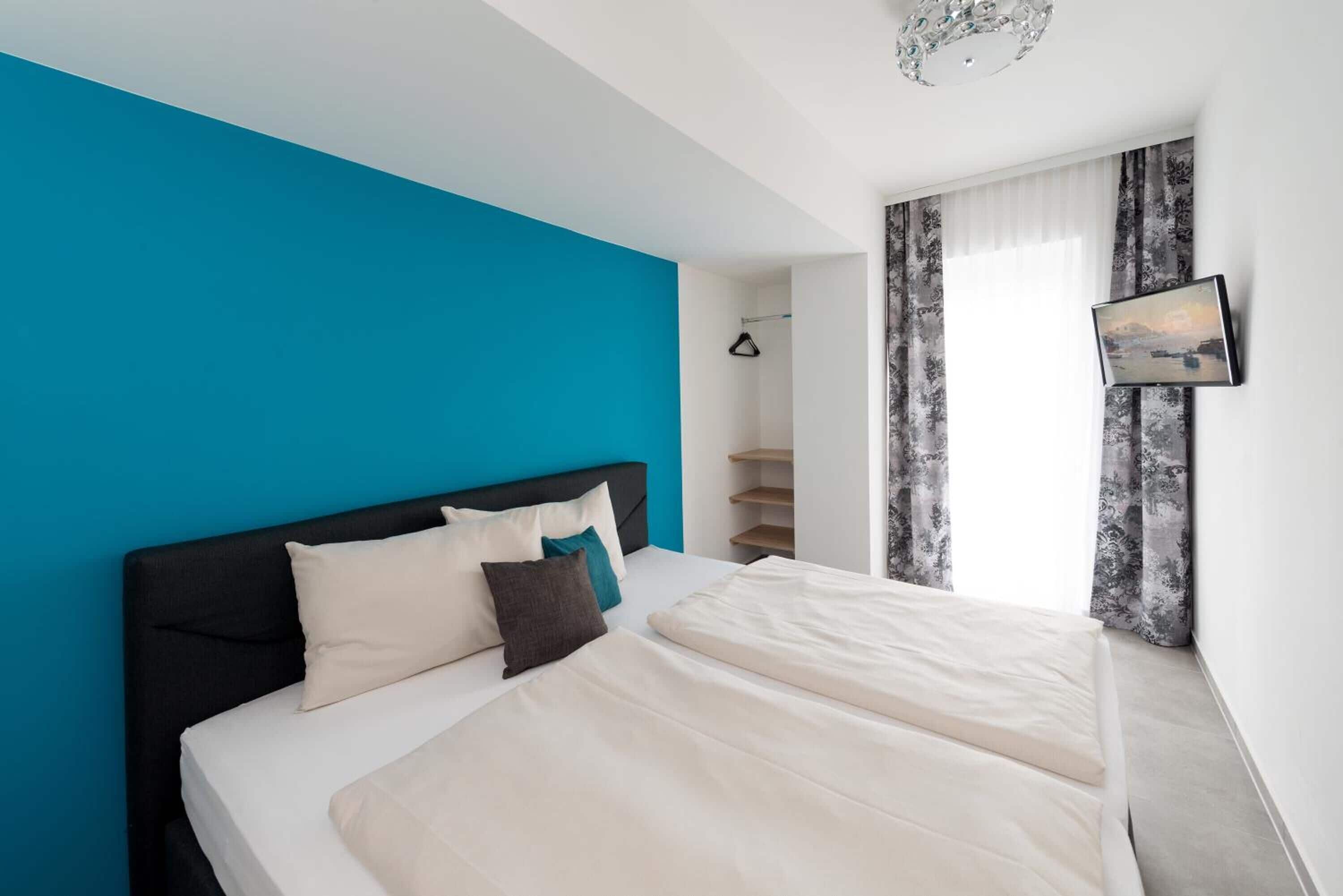 A bedroom with a large bed, a blue accent wall, a TV mounted on the wall, and a window with curtains.