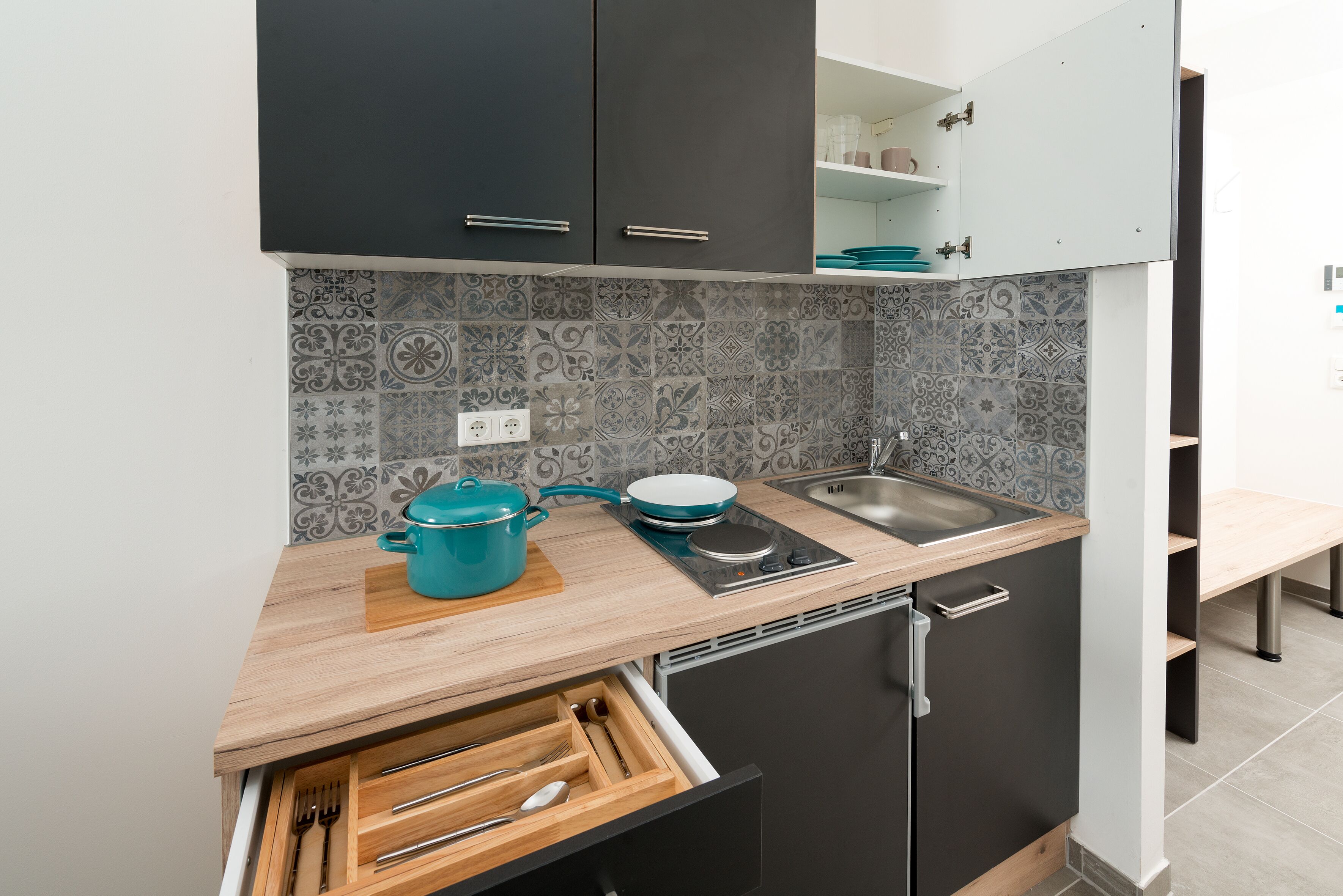 Cozinha compacta com armários escuros, bancada de madeira, pia e painel de azulejos estampados.