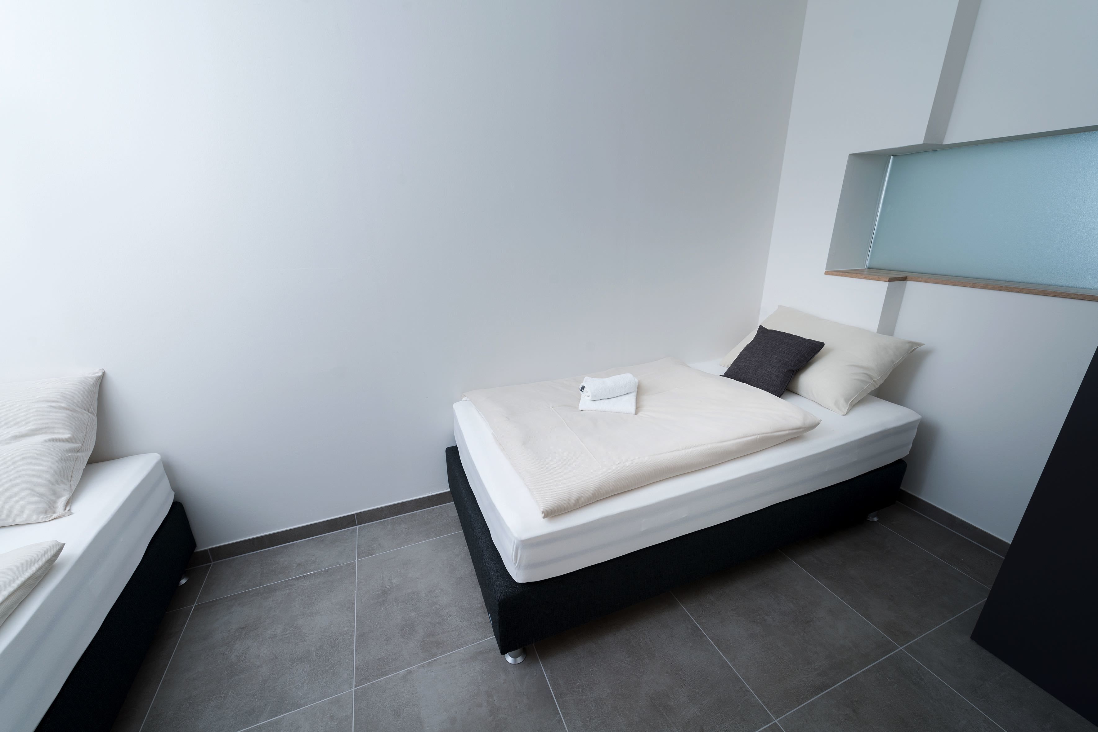 A small, modern bedroom with a single bed, a nightstand, and a built-in shelf above the bed.