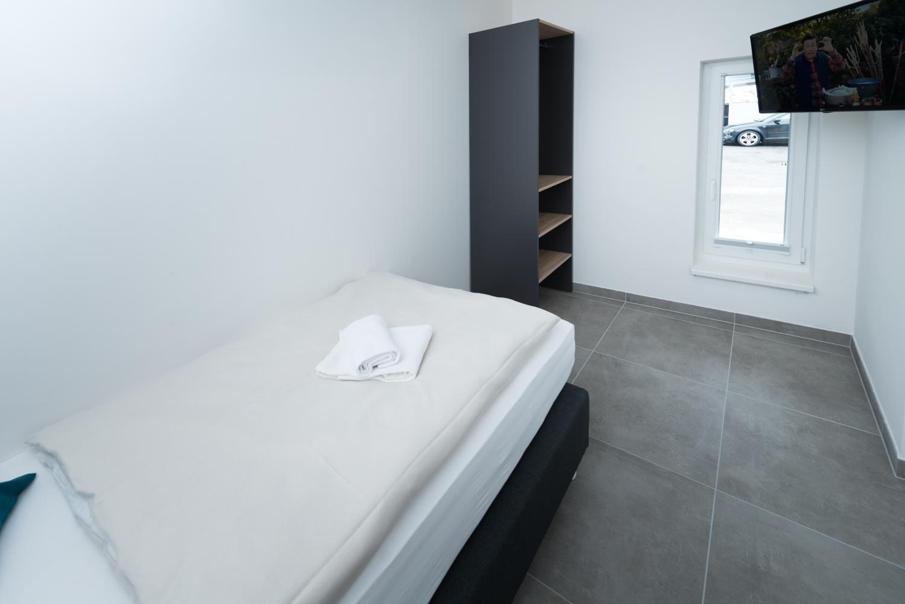 A single bed with a white mattress and a folded towel, a dark vertical storage unit, a television mounted above a window, and grey tiled flooring.