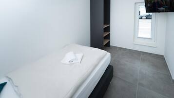 A single bed with a white mattress and a folded towel, a dark vertical storage unit, a television mounted above a window, and grey tiled flooring.