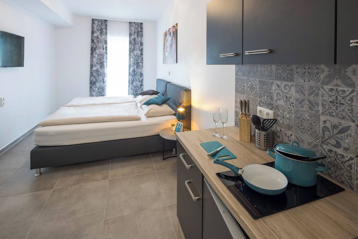 Ein kompaktes Hotelzimmer mit Bett, einem kleinen Nachttisch mit Lampe, einer Küchenzeile mit Herd und einer gefliesten Rückwand.