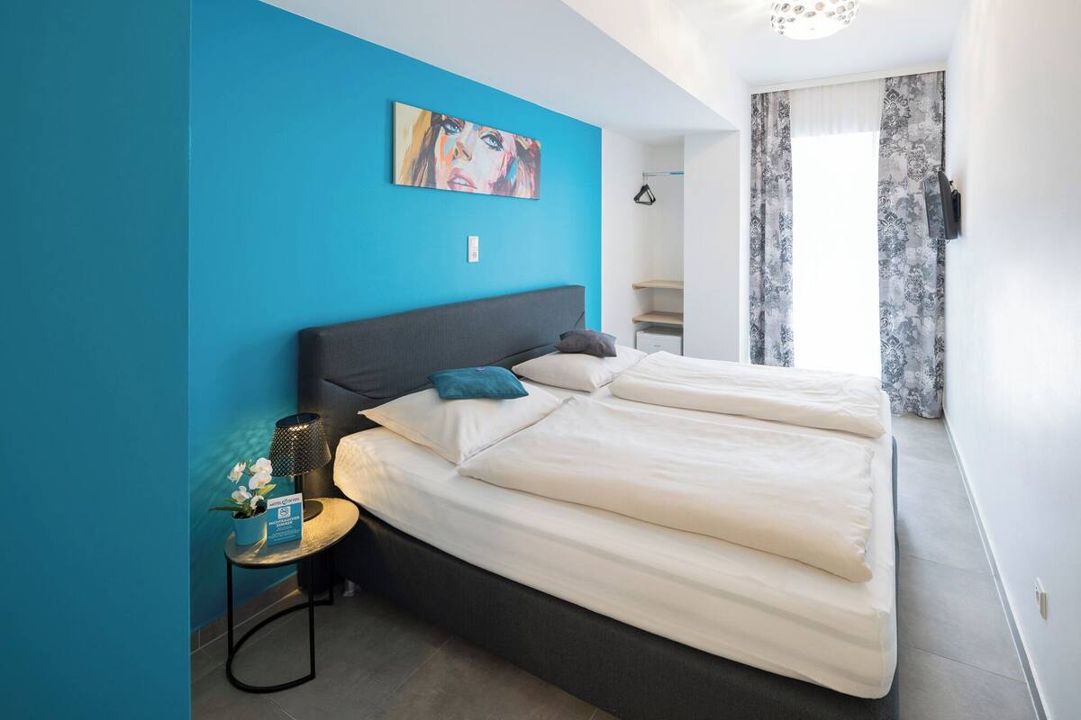 Ein modernes Hotelzimmer mit einem großen Bett, einem kleinen Nachttisch, einem Bild an der Wand und Blick ins Bad.