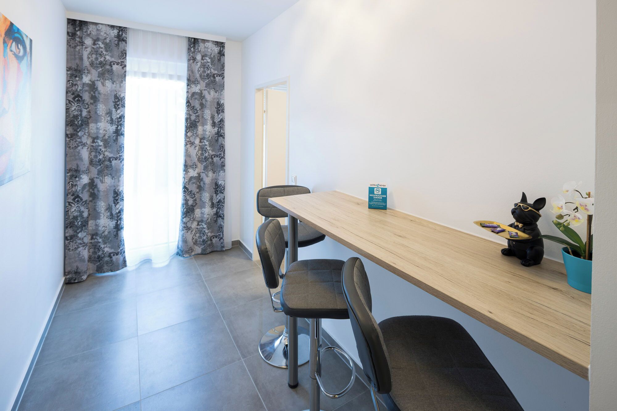 Una habitación de hotel moderna con una mesa de comedor de madera, sillas tapizadas en gris, una pequeña planta en maceta y un cartel que indica una política de no fumar.