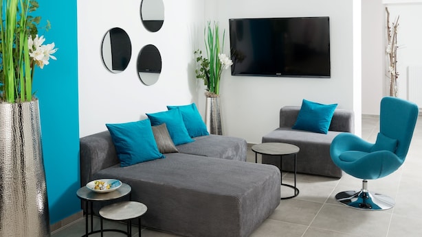 A modern living room with a grey sectional sofa, a blue armchair, two small round tables, and a flat-screen TV mounted on the wall. - Motel24seven (Bruck an der Mur)