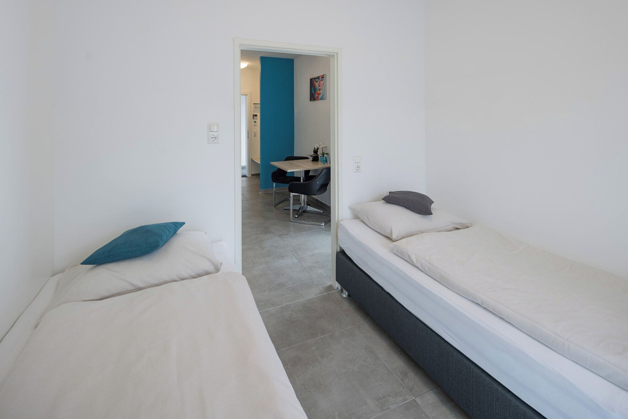 A narrow hallway with two single beds on either side, a small table with chairs in the background, and a blue accent wall.