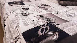 A bed with a newspaper print bedspread, a black headboard, and a folded towel on the side.