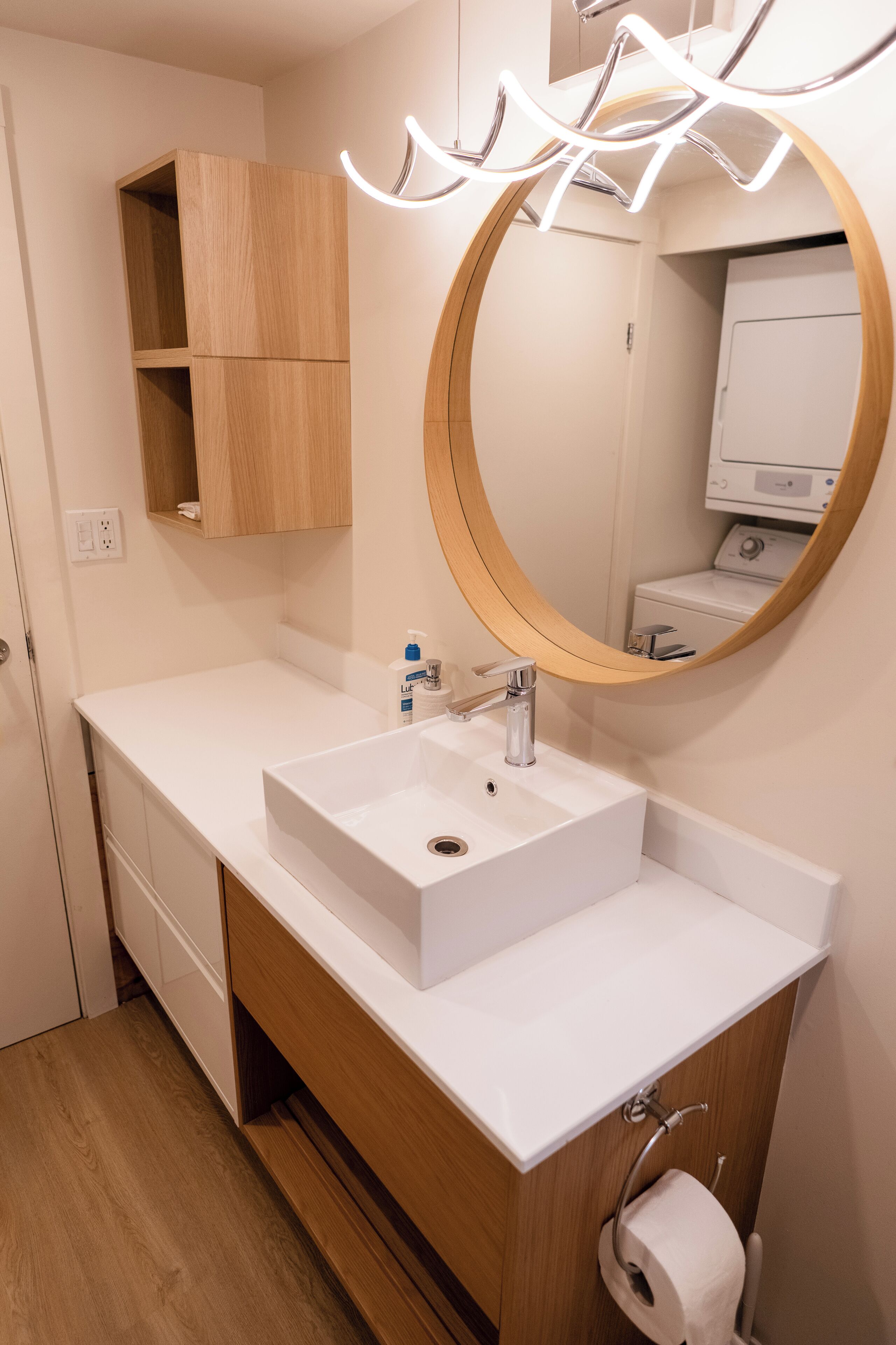 A modern bathroom with a white sink, wooden vanity, a round mirror, and a built-in washing machine.
